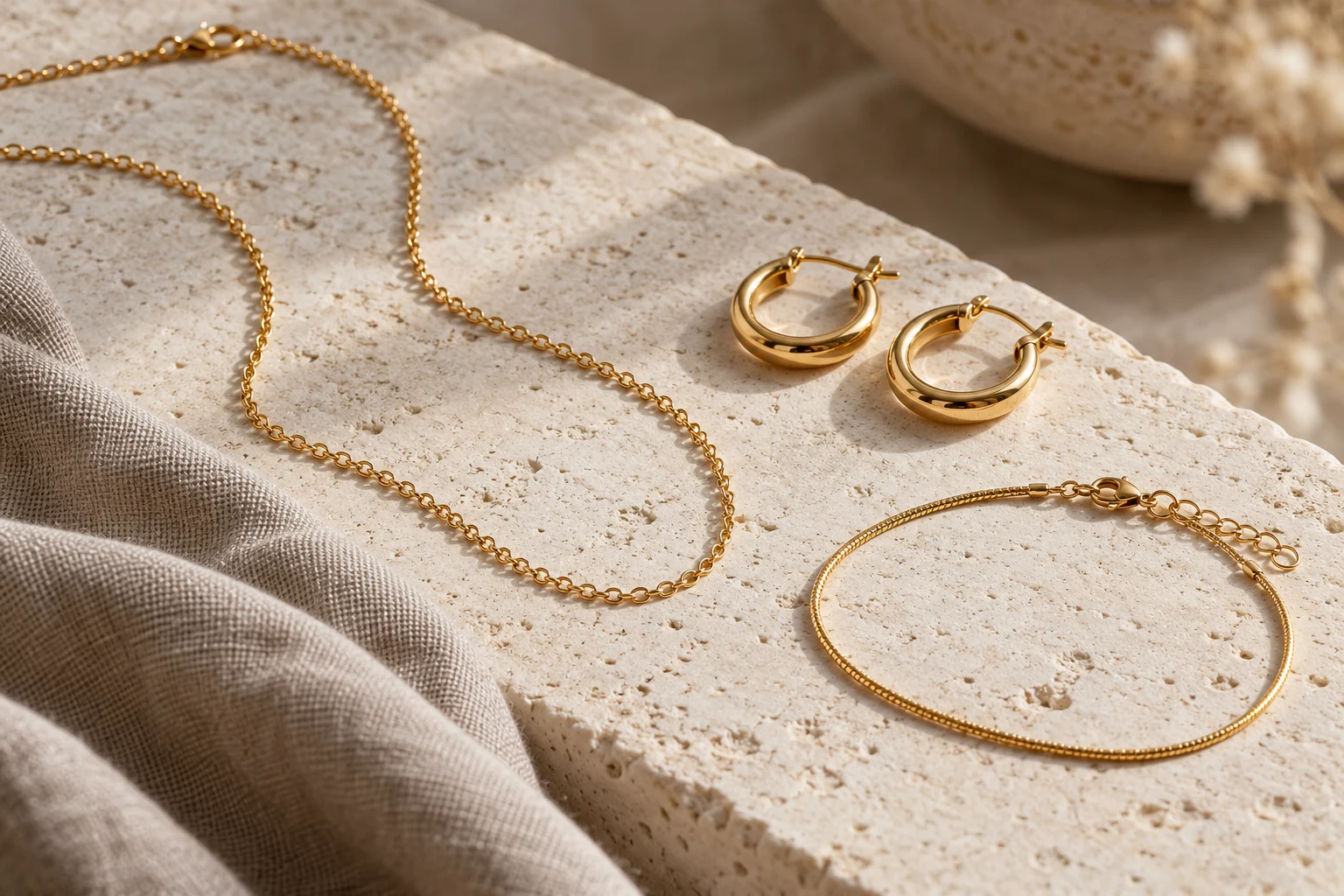 Gold-tone chain necklace, hoop earrings and bracelet arranged on textured cream stone beside taupe linen